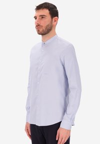 Uomo con capelli scuri e corti e barba, che indossa una camicia azzurro chiaro con colletto alla cinese e pantaloni neri, in piedi su uno sfondo bianco.