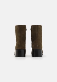 Botas de ante marrón hasta el tobillo con tacones cuadrados negros, que cuentan con una cremallera en la parte posterior. Textura suave y diseño simple y minimalista.