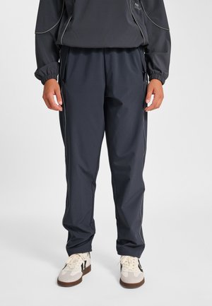 Mężczyzna ubrany w ciemnoszare spodnie sportowe z kieszeniami na zamek oraz białe tenisówki z czarnymi akcentami, stoi na tle jednolitego tła.