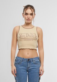 Šviesiai ruda sutrumpinta liemenė su ruda apdaila ir grafiniu „Von Dutch“ užrašu. Dėvimi lengvi mėlyni džinsai su šoniniu akcentu.