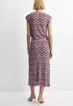 Donna con capelli biondi corti che indossa un vestito lavorato a maglia con motivo a chevron viola, marrone e bianco e sandali argento con tacco alto, vista di schiena.