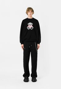 Sudadera negra con un gráfico de un oso de peluche gris, combinada con pantalones negros holgados y calzado negro texturizado. Diseño simple y casual.