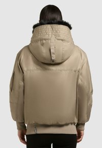 Giacca a vento beige con un grande cappuccio caratterizzato da un bordo in pelliccia nera, polsini elasticizzati e tasche laterali. Texture liscia e lucida.
