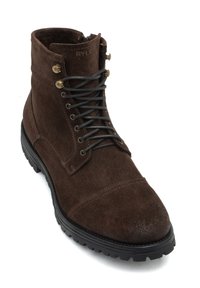 Stivale alla caviglia in suede marrone con suola in gomma nera, occhielli in metallo e lacci neri. Presenta dettagli di cucitura e una punta leggermente affusolata.