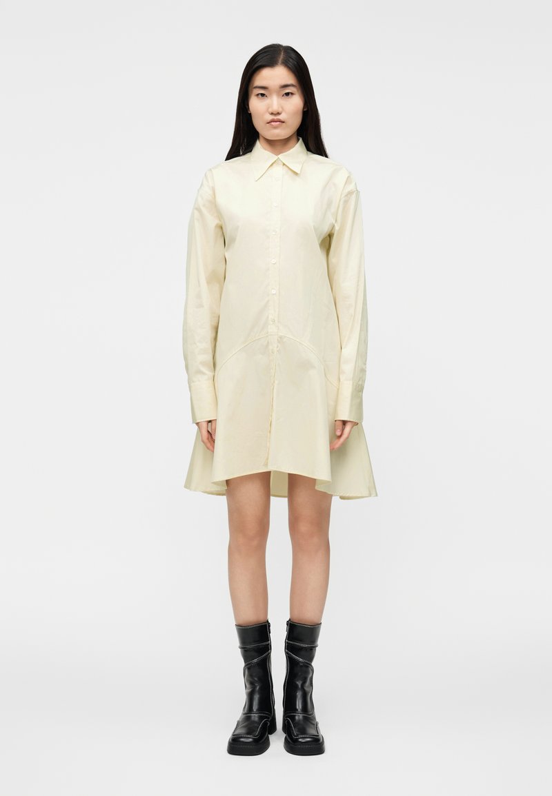 Šviesiai geltonos spalvos marškinių tipo suknelė su apykakle, ilgomis rankovėmis ir praplėstu apačios siluetu. Derinama su juodais, iki kelių siekiančiais, raišteliais surišamais batais.