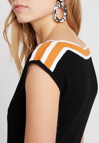 Žena v černém bezramínkovém topu s oranžovými a bílými pruhy na ramenech a černo-bílými oválnými náušnicemi se vzorem.