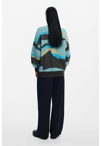 Maglione lavorato a maglia oversize con motivi astratti di montagne multicolori in turchese, giallo, grigio e marrone scuro. Indossato con pantaloni a gamba larga.
