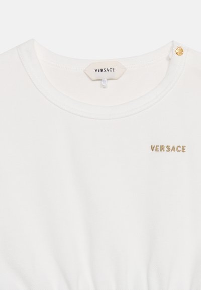Λευκό πουκάμισο Versace με χρυσό κεντημένο λογότυπο στο στήθος, στρογγυλή λαιμόκοψη και λεπτομέρεια με χρυσό κουμπί στον ώμο.