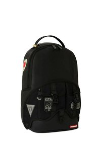Mochila negra hecha de un tejido duradero, con un logotipo redondo en rojo y blanco, bolsillos con cremallera en la parte frontal y varios parches. Textura suave.