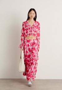 Completo floreale a due pezzi con top corto arricciato e gonna lunga in tessuto rosa con motivi di fiori rossi e verdi; abbinato a sandali azzurri.
