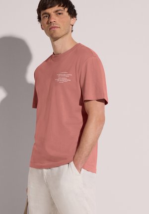 Jeune homme portant un T-shirt rose poudré avec un texte blanc sur la poitrine et un pantalon beige clair, debout, une main dans la poche, devant un fond clair.