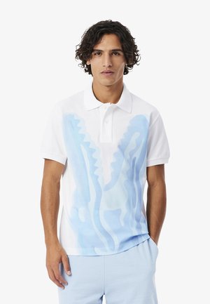 Jeune homme aux cheveux bouclés mi-longs portant un polo blanc avec un motif abstrait bleu clair et un pantalon bleu clair.