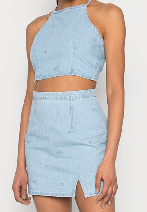 Kobieta nosząca jasnoniebieski jeansowy crop top z wiązaniem na szyi i pasującą mini spódnicę z wysokim stanem i małym rozcięciem z przodu.