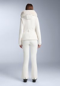 Jachetă albă cu glugă din blană sintetică, talie încinsă și design ajustat, asortată cu pantaloni albi evazați cu fermoare la tivuri. Textură fină.