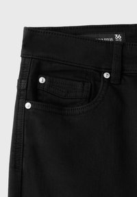 Gros plan sur la ceinture et la poche avant d'un jean en denim noir avec rivets métalliques et étiquette de taille visible 36 USA 04.