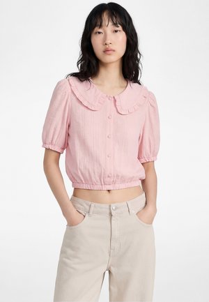 Femme aux cheveux noirs portant un chemisier rose clair à manches courtes avec col à volants et pantalon beige, mains dans les poches, fond neutre.