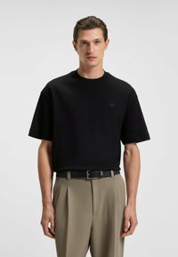 Zwarte korte mouwen t-shirt met een ronde halslijn, met een subtiele logo op de borst. Gecombineerd met lichtbeige geplooide broek en een zwarte riem.