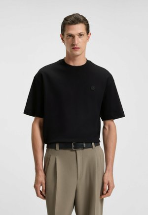Maglietta nera a maniche corte con scollo rotondo, con un logo discreto sul petto. Abbinata a pantaloni beige chiaro con pieghe e una cintura nera.