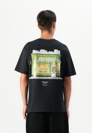 Man with short hair wearing a black t-shirt showing a green storefront illustration labeled "FRANKS" on the back.