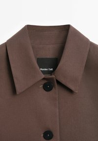 Hnědá košile s špičatým límcem, která má černé knoflíky a tkanou texturu. Štítek nese nápis "Massimo Dutti" na černém tagu.