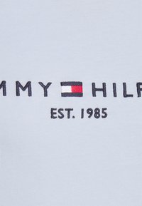 Vaaleansininen puuvillakangas, jossa on brodeerattu "TOMMY HILFIGER" -logo ja vaakasuora väri-iloittelu punaisessa, valkoisessa ja laivastonsinisessä.