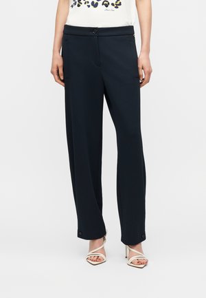Woman wearing straight-leg black pants with button details at cuffs and white strappy high-heeled sandals on a white background.