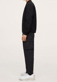 Veste bomber noire avec poignets côtelés, associée  à un pantalon cargo noir avec de grandes poches latérales. Complété par des baskets blanches.