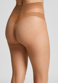 Nahaufnahme einer Person, die transparente beige Strumpfhosen mit verstärktem Oberschenkel- und Taillenbund vor einem schlichten Hintergrund trägt.