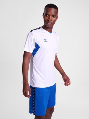 Mann steht, trägt weißes Sporttrikot mit blauen Akzenten und blaue Shorts mit schwarzen Chevron-Streifen an der Seite, schaut nach rechts.