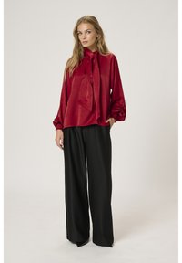 Blusa rossa di raso con un nodo al collo e maniche lunghe e arricciate, abbinata a pantaloni neri a gamba larga in un tessuto liscio e leggero.