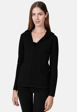 Sweat à zip noir en matériau doux, avec un design ajusté, des poches et une capuche, associé à un simple haut noir et à un jean.