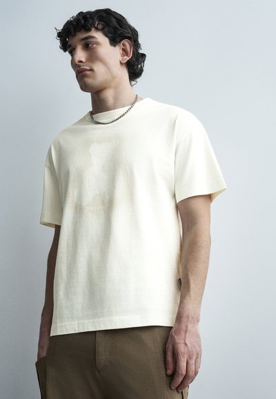Junger Mann mit kurzen lockigen Haaren trägt ein cremefarbenes T-Shirt mit Teddybär-Motiv, braune Hose, silberne Kettenkette und einen Steckerohrring, steht vor einer grauen Wand.