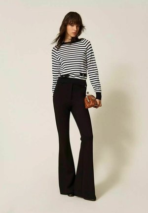 Donna che indossa una maglia a maniche lunghe a righe bianche e nere, pantaloni neri a vita alta a zampa, che tiene una piccola borsa marrone trapuntata.