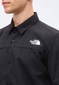 Άνδρας φοράει μαύρο πουκάμισο με κουμπιά και γιακά, με το λογότυπο "The North Face" στο αριστερό μέρος του στήθους.