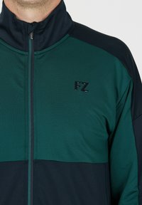 Zip-up jacka med en mörkgrön kropp och marinblå detaljer. Har en hög krage och frontdragkedja. Svart "FZ"-logotyp på bröstet.