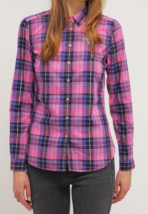 Femme portant une chemise ajustée à carreaux rose et bleue à manches longues boutonnée et un jean foncé, debout devant un fond uni.