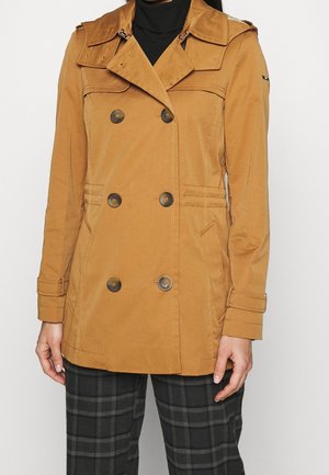 Femme portant un trench-coat croisé couleur camel avec des détails de boutons et un pantalon noir à carreaux.