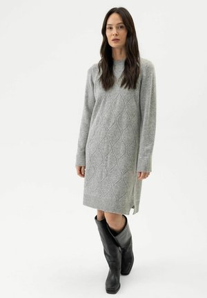 Frau in einem grauen, kabelgestrickten Pulloverkleid und schwarzen kniehohen Stiefeln steht vor einem schlichten weißen Hintergrund.