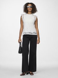 Femeie purtând ochelari de soare negri, top fără mâneci din dantelă albă, blugi largi negri, sandale cu toc negre, ținând o geantă mică neagră.