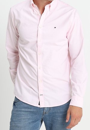 Homme portant une chemise rose pâle à boutons avec manches longues et un jean bleu, debout avec une main dans la poche.
