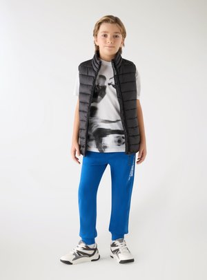 Ragazzo in piedi che indossa un gilet imbottito nero, una t-shirt bianca con stampa grafica, pantaloni da jogging blu e sneaker bianche su sfondo semplice.