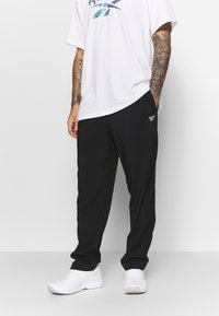 Calças de desporto pretas feitas de um tecido leve, com bolsos e um logo da Reebok. Combinadas com uma t-shirt branca larga. Ténis brancos.