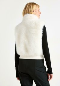 Valkoinen tekoturkisliivi ribatulla helmalla, käytössä mustan pitkähihainen paidan päällä. Sileä rakenne ja minimalistinen muotoilu, suunniteltu kerroksellista tyyliä varten.