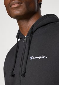 Schwarzer Kapuzenpullover mit Frontreißverschluss, Zugbändern mit strukturierten Enden und einem weißen gestickten "Champion"-Logo auf der Brust.