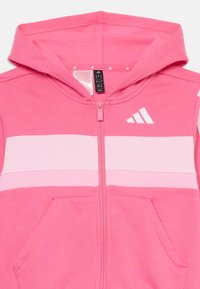 Haut de sweatshirt zippé rose avec un design contrasté intégrant des rayures rose clair, des poches kangourou et un logo Adidas blanc sur la poitrine.
