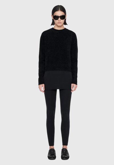 Un mannequin se tient devant un fond gris clair, portant des lunettes de soleil noires, un pull noir, un legging et des baskets.