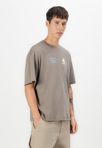 Young man with short dark hair wearing a loose gray t-shirt with text and beige pants, standing against a plain white background.