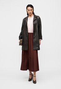 Femme portant une veste en cuir noire, un chemisier rose clair, une jupe large marron, des talons noirs, tenant une pochette à motif léopard sur un fond uni.