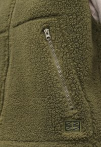 Flisasta brezha v olivno zeleni barvi, z žepom na zadrgo in teksturirano površino. Vključuje majhno, našito oznako z geometrijskim logotipom.