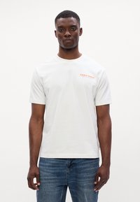 Λευκό βαμβακερό t-shirt με κοντά μανίκια, που φέρει το κείμενο "EARLY BIRDS" σε πορτοκαλί χρώμα στο αριστερό στήθος. Φοριέται με μπλε τζιν παντελόνι.